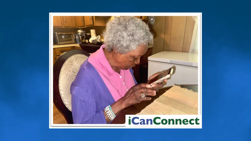 Side profile of an elderly woman seated at a table, holding a smartphone in both hands while closely looking down at the screen