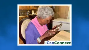 Side profile of an elderly woman seated at a table, holding a smartphone in both hands while closely looking down at the screen